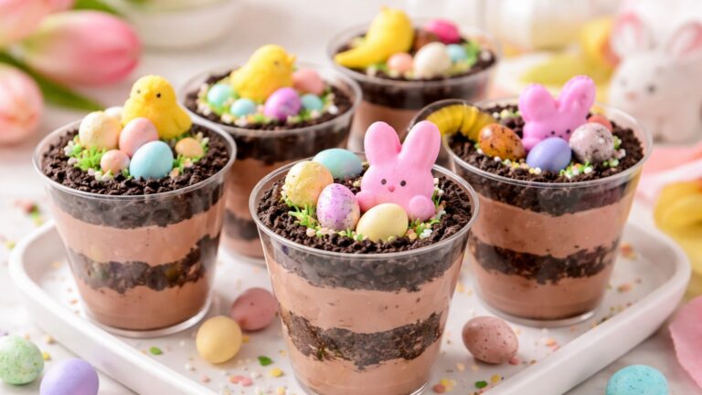 Easter dirt cups layered with chocolate pudding and crushed cookies in clear cups, topped with pastel mini eggs, marshmallow chicks and bunnies, gummy worms, and spring sprinkles on a light Easter table setting.