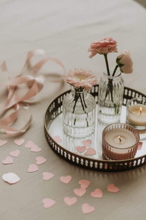 A close-up flat lay of Galentine’s Day decor details including heart confetti, mini glass vases, candles, blush ribbons, and soft pink accents.