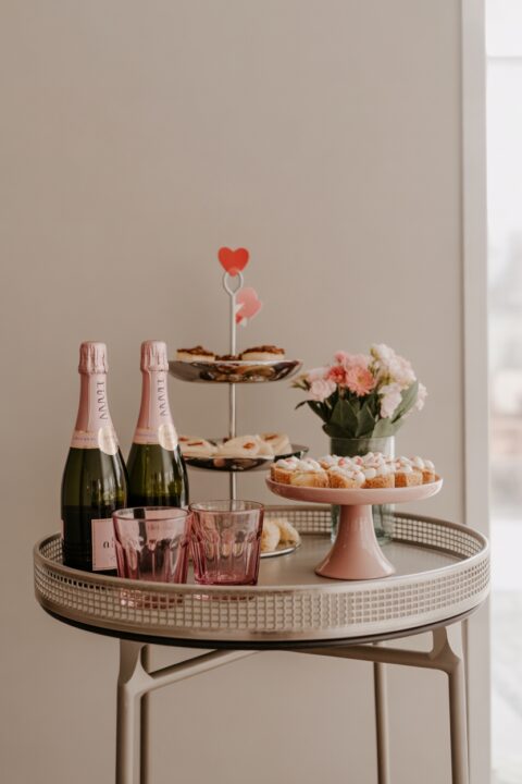 A chic Galentine’s Day drink and snack station with pink glassware, decorative tray, desserts, flowers, and feminine decor accents on a side table.