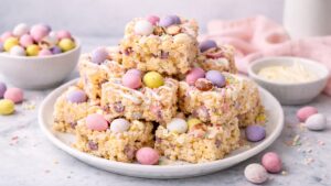 A plate of Mini Egg Rice Krispie Treats stacked in neat squares, topped with pastel Mini Eggs and white chocolate drizzle, styled on a light kitchen surface with soft spring colours in the background