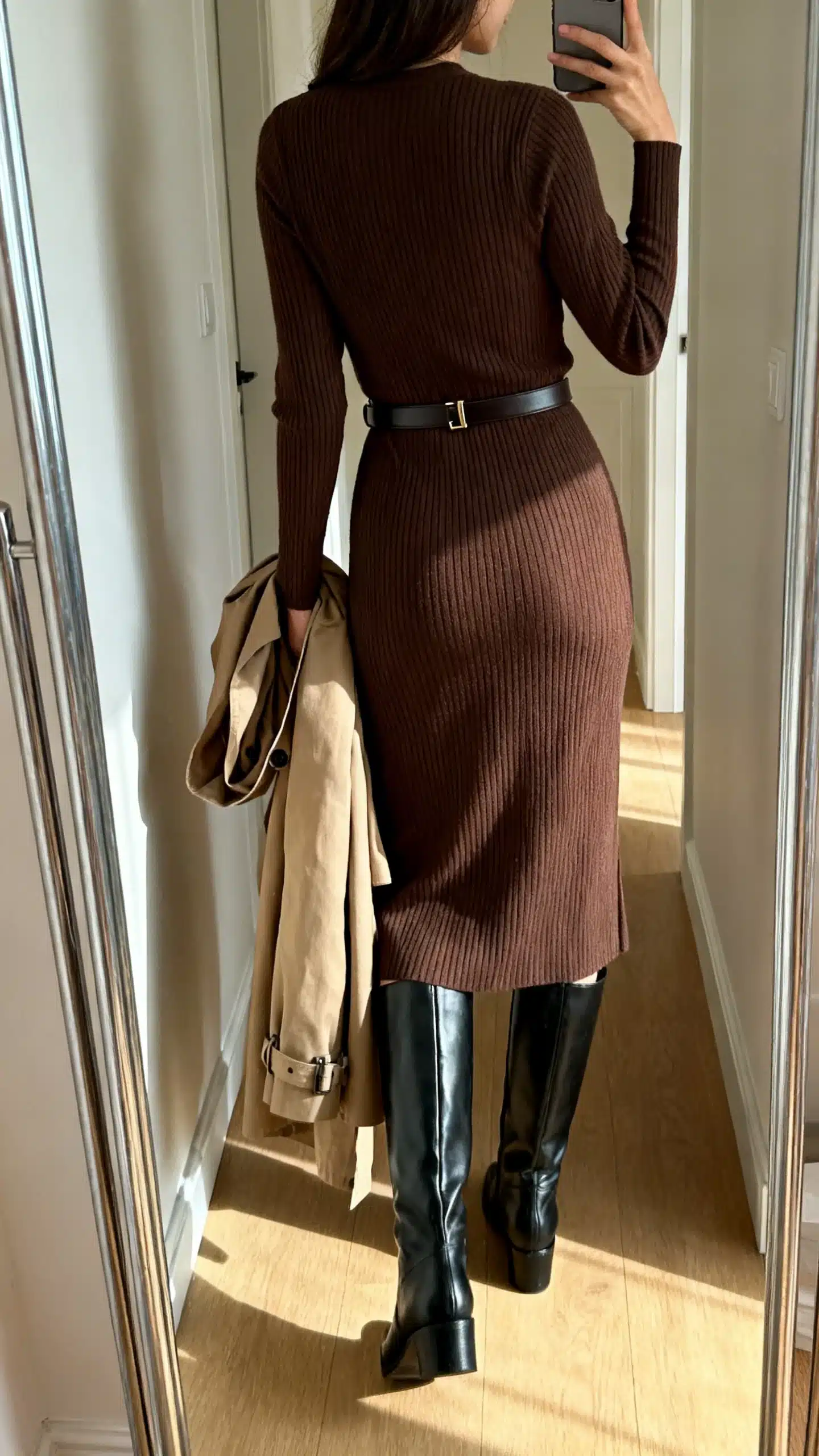 iPhone back-view hallway mirror selfie of a woman in a ribbed knit midi dress in chocolate with black knee-high boots, slim belt, and a trench draped over her arm, face not visible, natural afternoon light, iPhone photo quality.