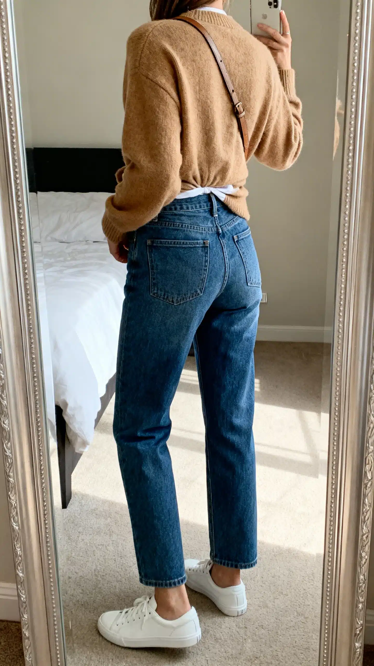 iPhone back-view mirror selfie of a woman wearing straight-leg blue jeans with a camel cashmere sweater half-tucked, white low-top sneakers, and a crossbody bag, face not visible, bedroom mirror, natural daylight, iPhone photo quality