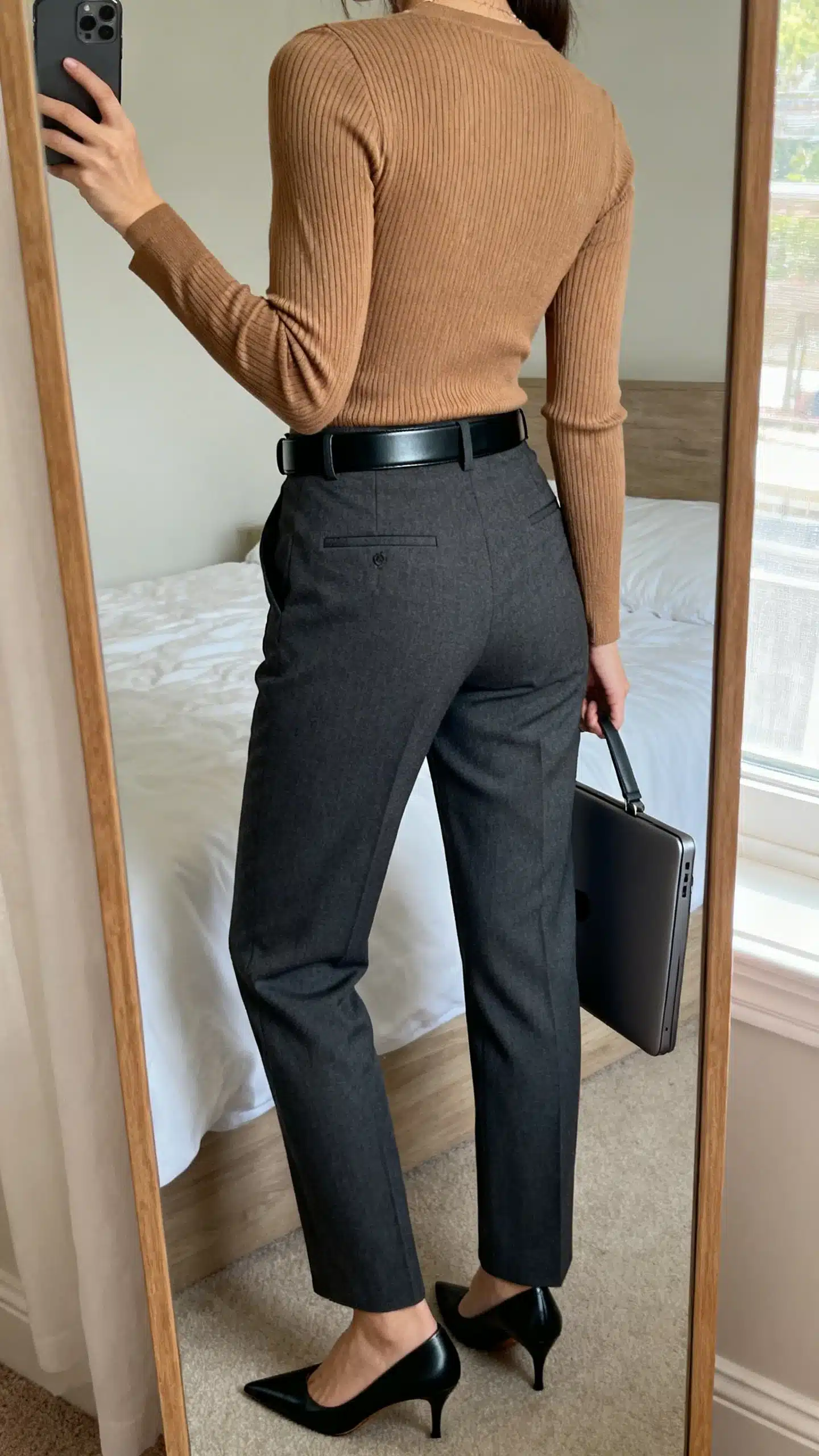 iPhone back-view mirror selfie of a woman wearing tailored charcoal trousers with a fitted ribbed knit top in camel, sleek black belt, pointed toe pumps, and a structured laptop bag, face not visible, bedroom mirror, soft window light, iPhone photo quality.