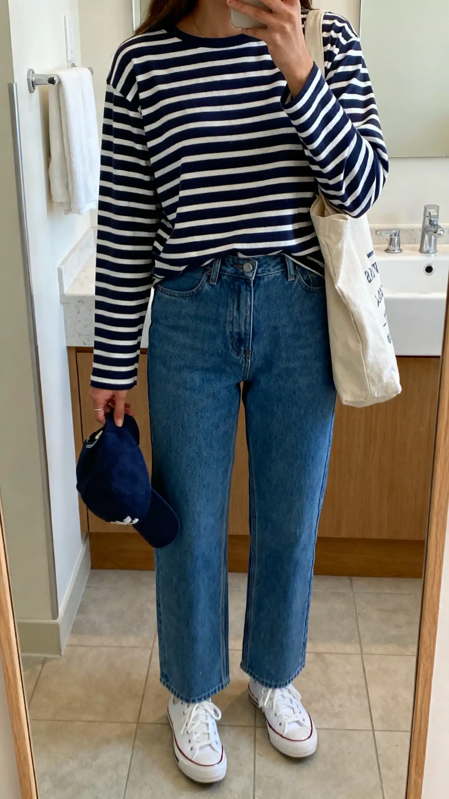 iPhone casual mirror selfie of a woman wearing a navy-and-white Breton striped long-sleeve tee with relaxed straight blue denim, white canvas sneakers, baseball cap in hand, and a canvas tote, face not visible, cropped at neck, bathroom mirror, indoor lighting, iPhone photo quality.