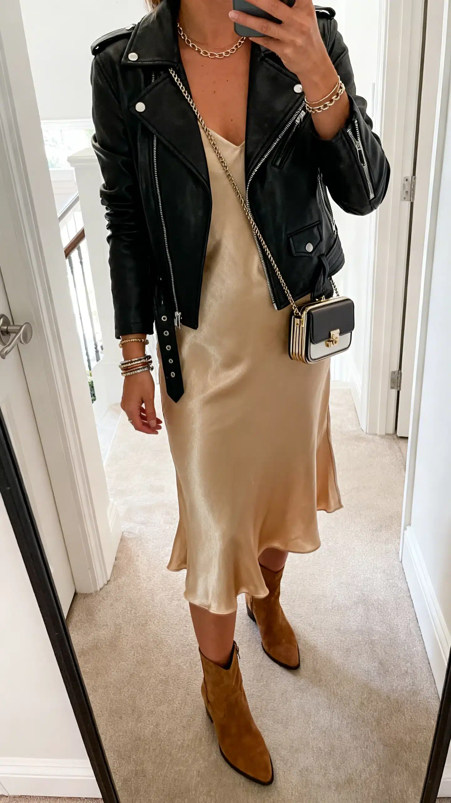 iPhone mirror selfie of a woman in a black leather moto jacket over a champagne slip dress, ankle boots, a compact crossbody, delicate chain necklace, and stacked bracelets, face not visible, cropped at shoulders, entryway mirror, natural daylight, iPhone photo quality.