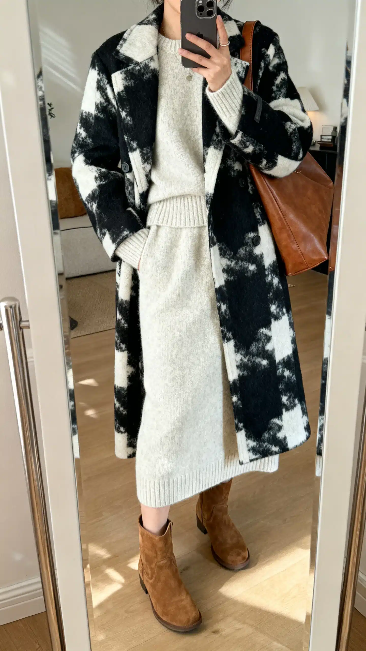 iPhone mirror selfie of a woman in a bold statement coat layered over a monochrome knit set (matching sweater and knit skirt), suede ankle boots, and a leather shoulder bag, face not visible, cropped at shoulders, full-length mirror, cozy indoor lighting, iPhone photo quality