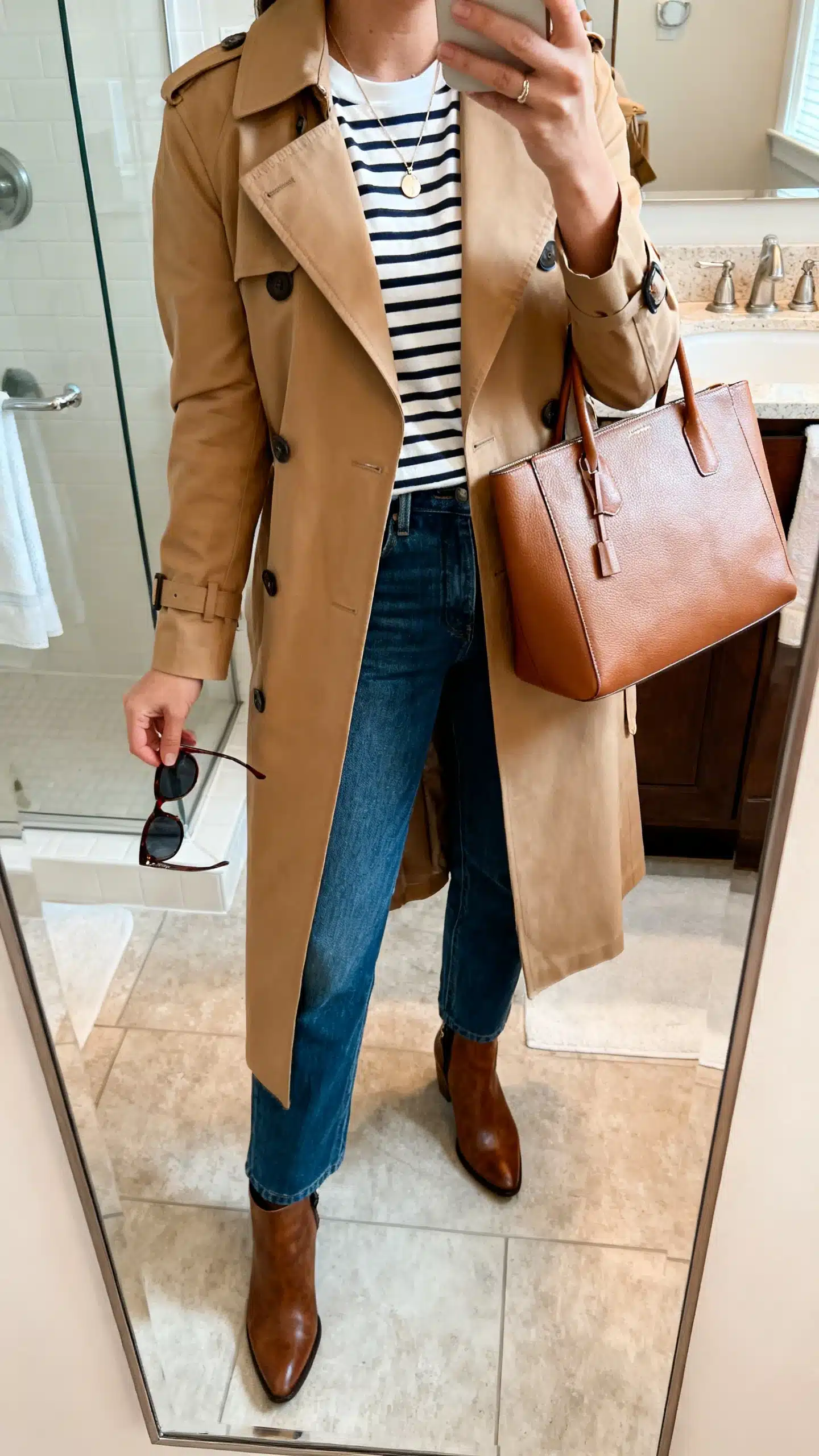 iPhone mirror selfie of a woman in a camel mid-length trench coat over a striped Breton tee with straight-leg jeans, ankle boots, a structured leather tote, delicate pendant necklace, and sunglasses in hand, face not visible, bathroom mirror, soft indoor morning light, iPhone photo quality.