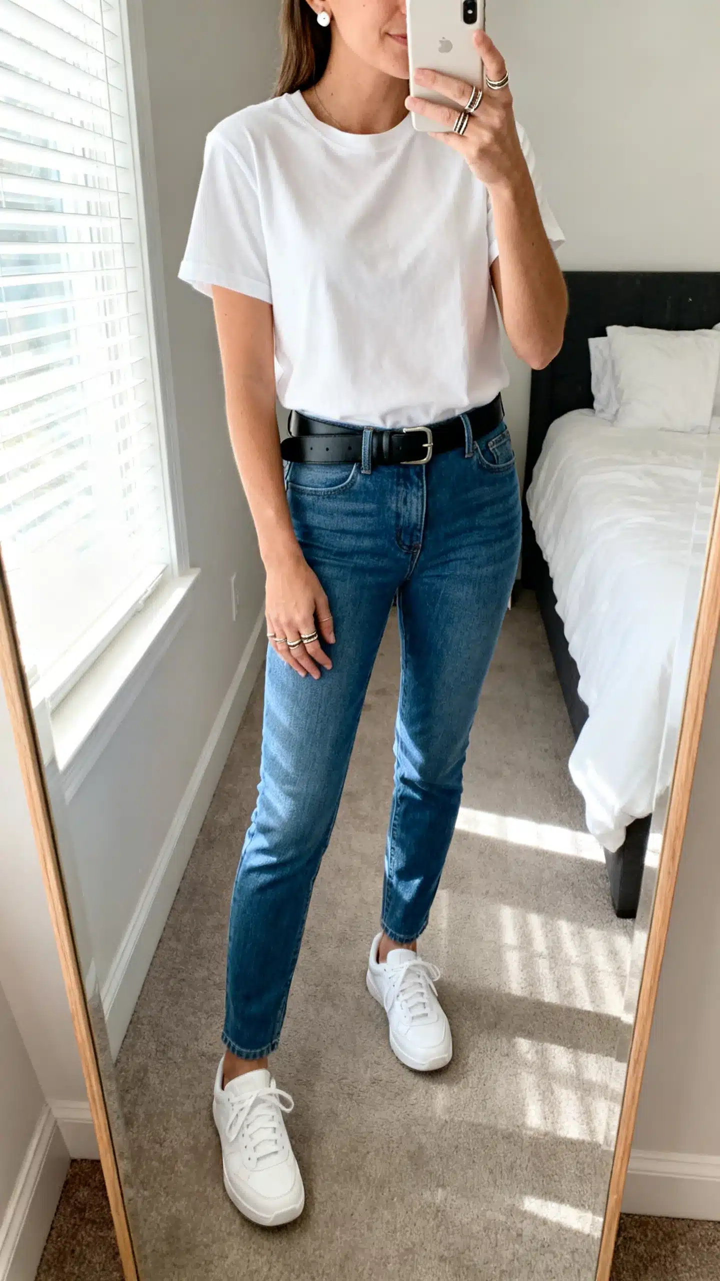 iPhone mirror selfie of a woman in a high-quality white T-shirt tucked into mid-blue slim-straight jeans, clean white sneakers, a black leather belt, everyday stud earrings, and stackable rings, face not visible, cropped at neck, bedroom mirror, bright window light, iPhone photo quality.