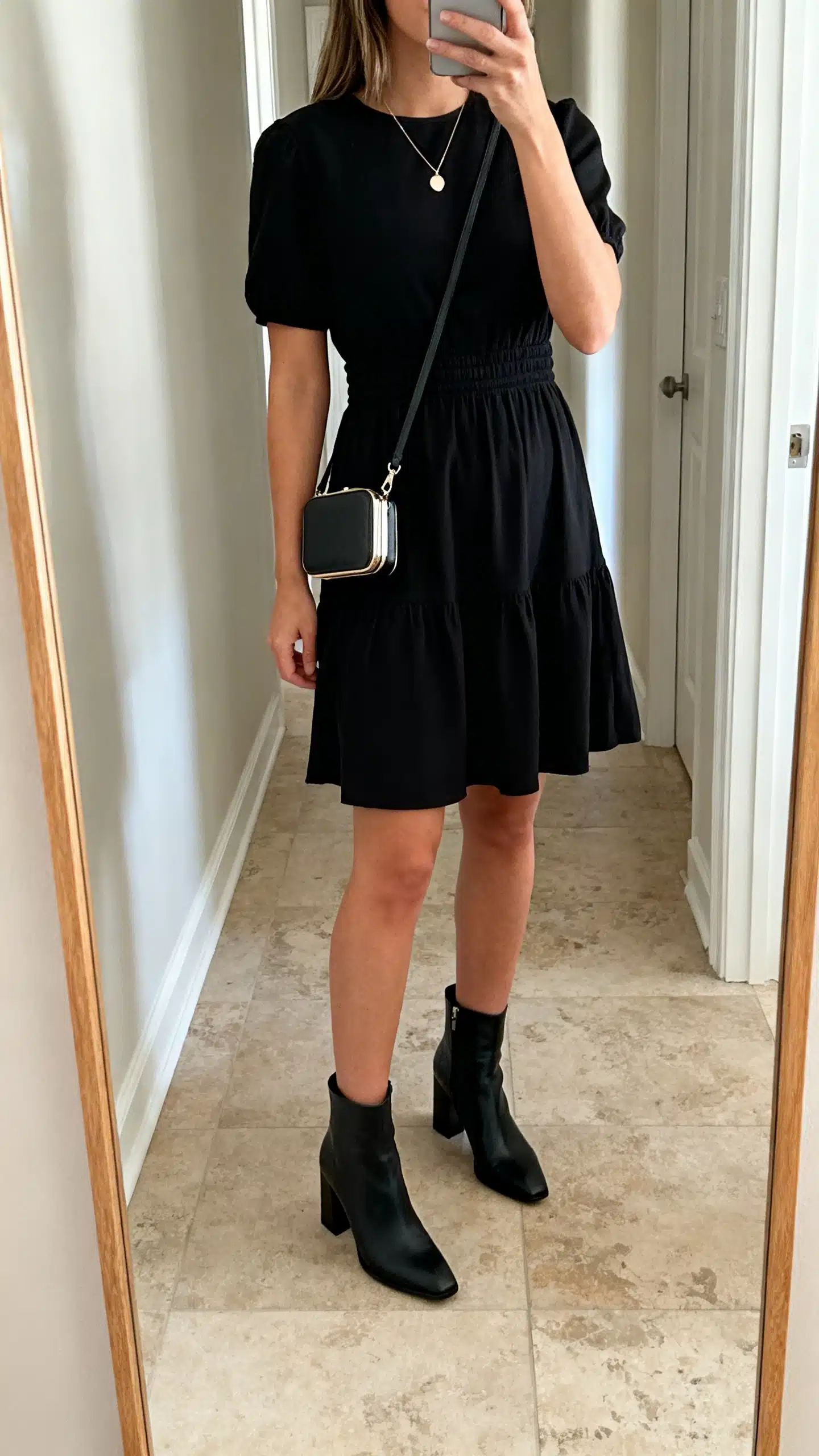 iPhone mirror selfie of a woman in a little black dress with a defined waist, black heeled ankle boots, delicate pendant necklace, and a compact crossbody bag, face not visible, cropped at shoulders, hallway full-length mirror, indoor lighting, iPhone photo quality.