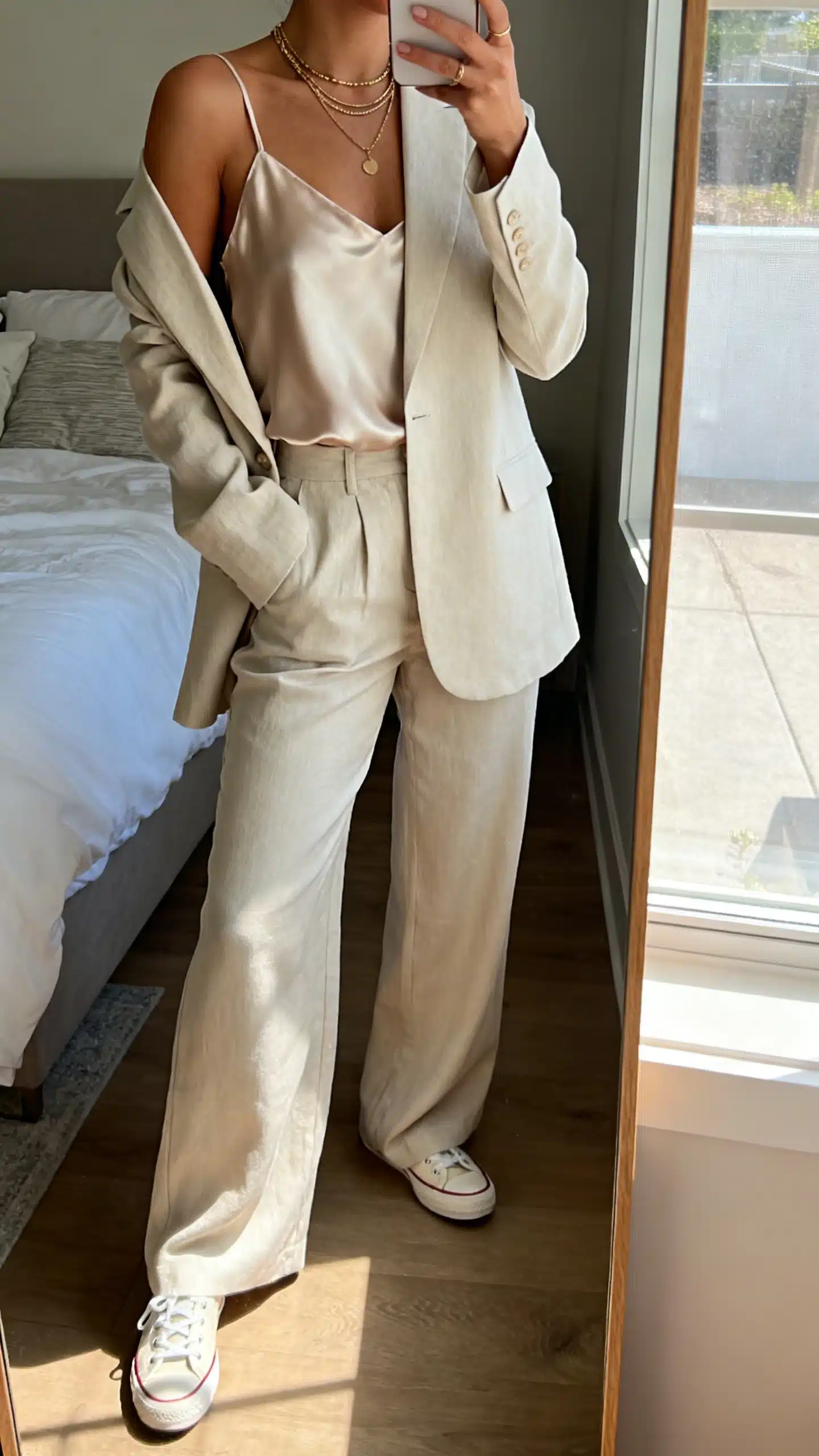 iPhone mirror selfie of a woman in a relaxed suit set (soft-shoulder blazer and matching wide-leg trousers) with a silk camisole, sleek sneakers, and layered necklaces, face not visible, bedroom mirror, morning window light, iPhone photo quality