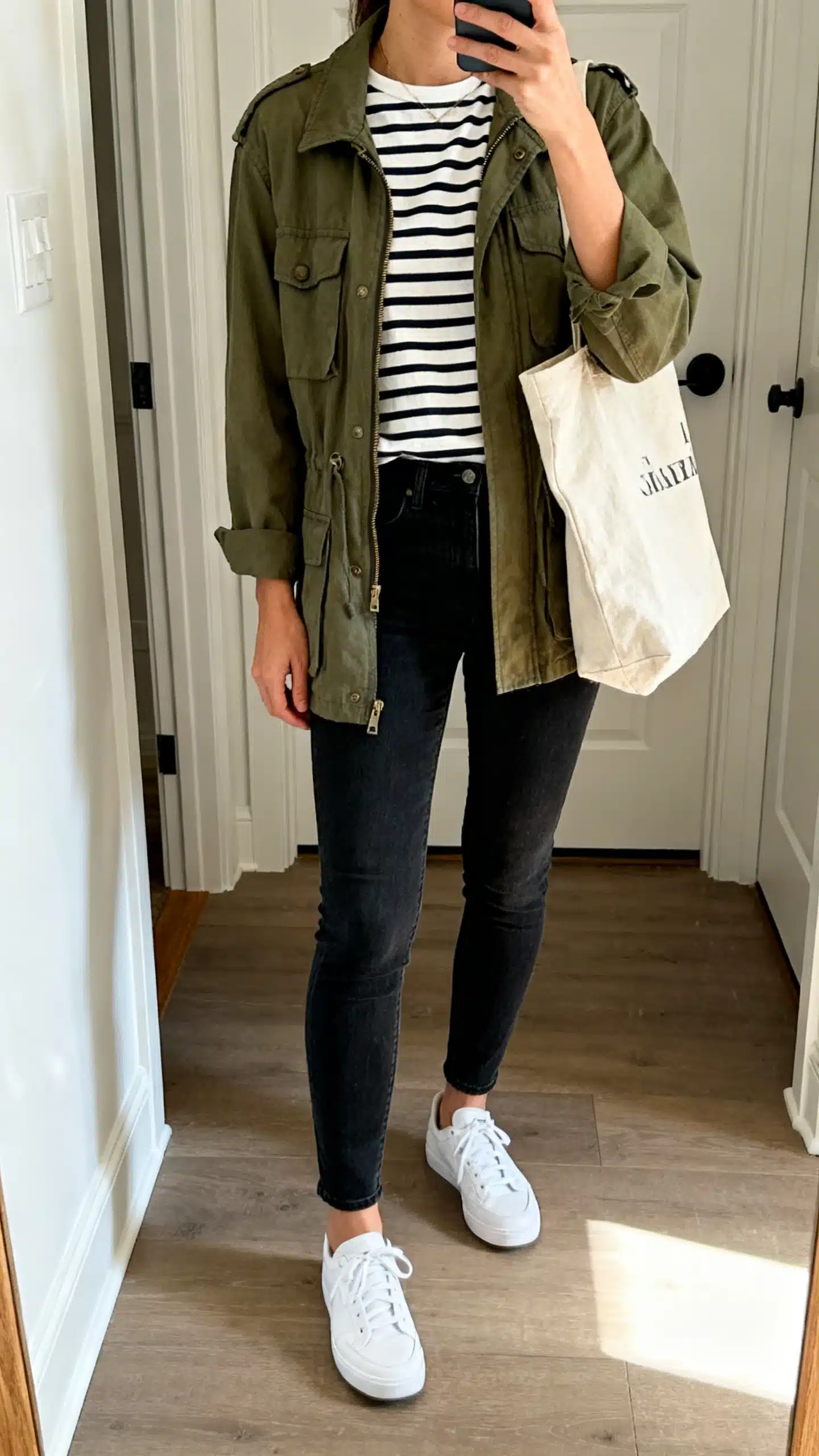 iPhone mirror selfie of a woman wearing a Breton striped top, olive utility jacket, slim dark jeans, and clean white sneakers, with a canvas tote, face not visible, cropped at neck, entryway mirror, natural daylight, iPhone photo quality