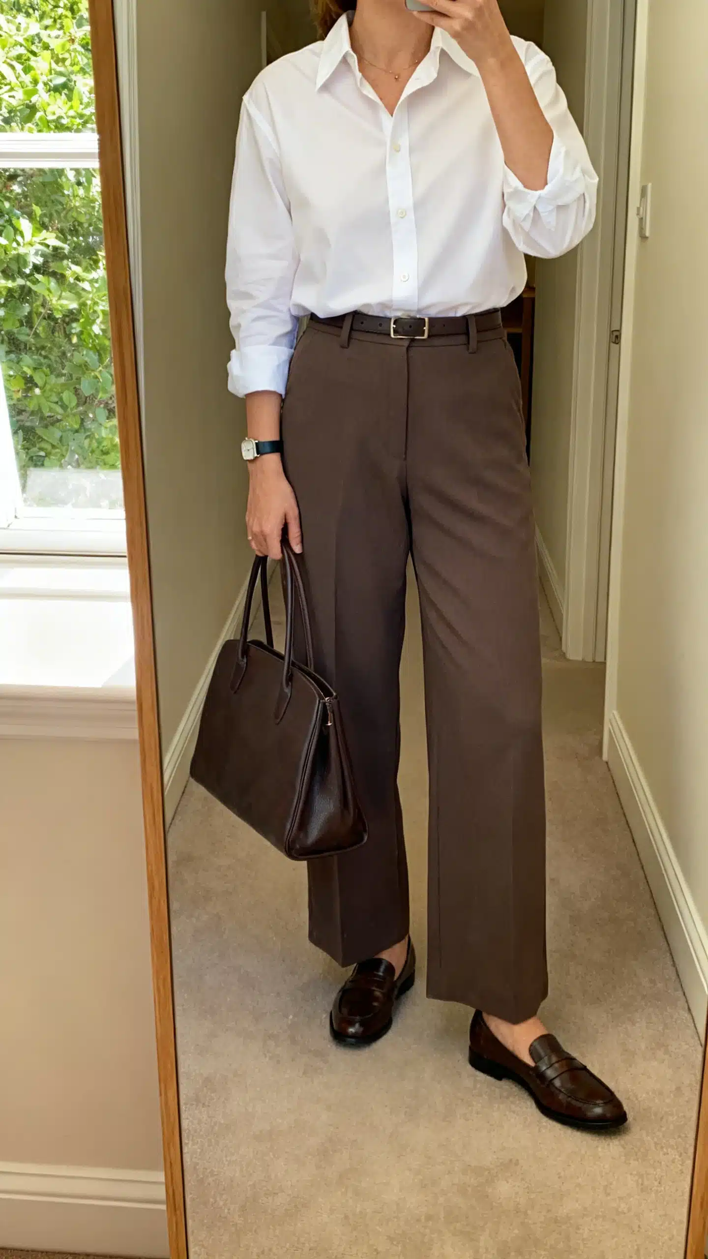 iPhone mirror selfie of a woman wearing a crisp button-down shirt tucked into wide-leg trousers, classic loafers, structured tote, slim watch, face not visible, cropped at shoulders, hallway mirror, natural daylight from window, iPhone photo quality