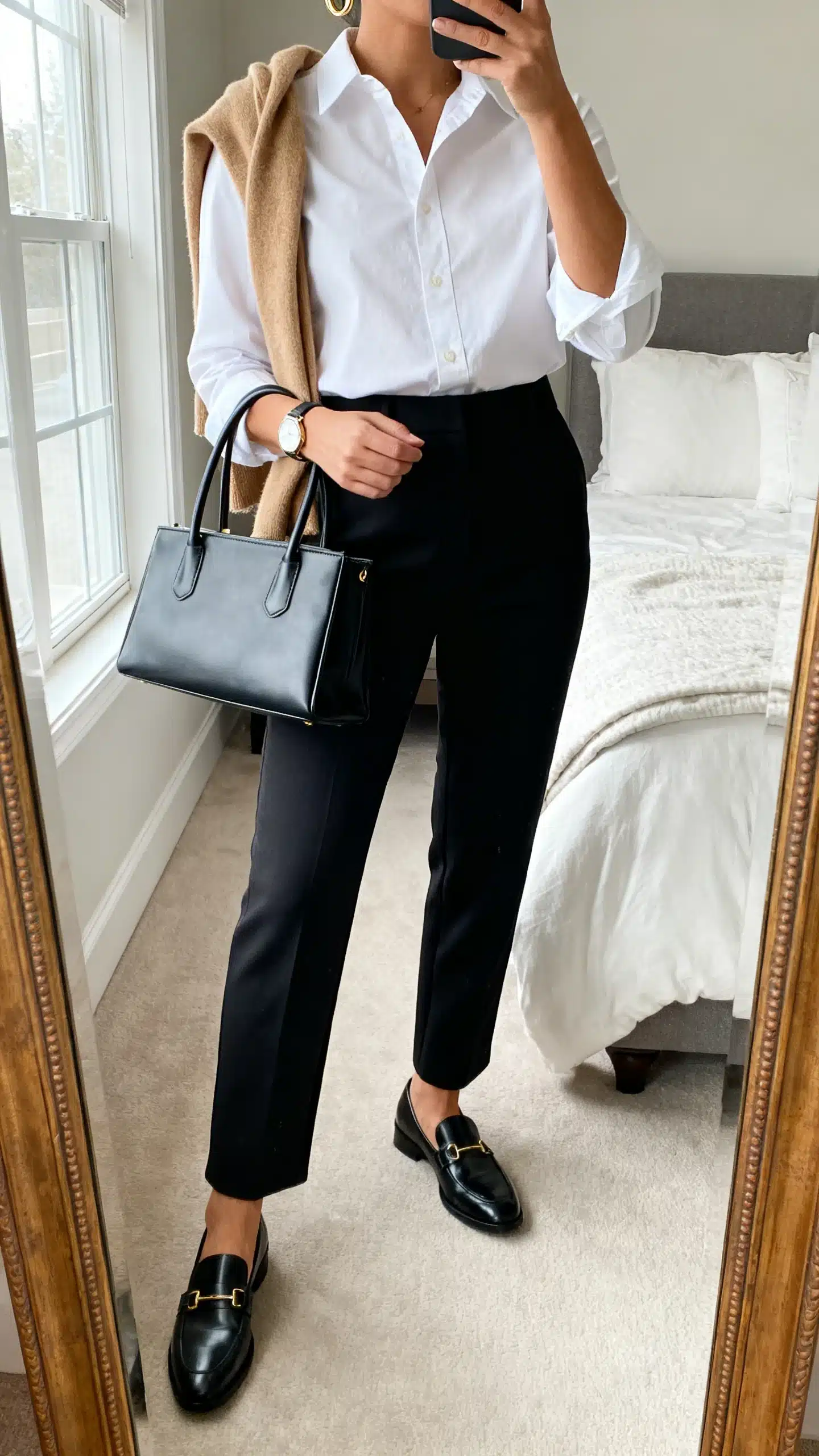 iPhone mirror selfie of a woman wearing a crisp white button-down tucked into high-waisted black tailored trousers, polished black loafers, a structured top-handle bag, minimal gold hoops, and a simple watch; optional camel cashmere draped over shoulders, face not visible, cropped at neck, bedroom mirror, natural window light, iPhone photo quality.