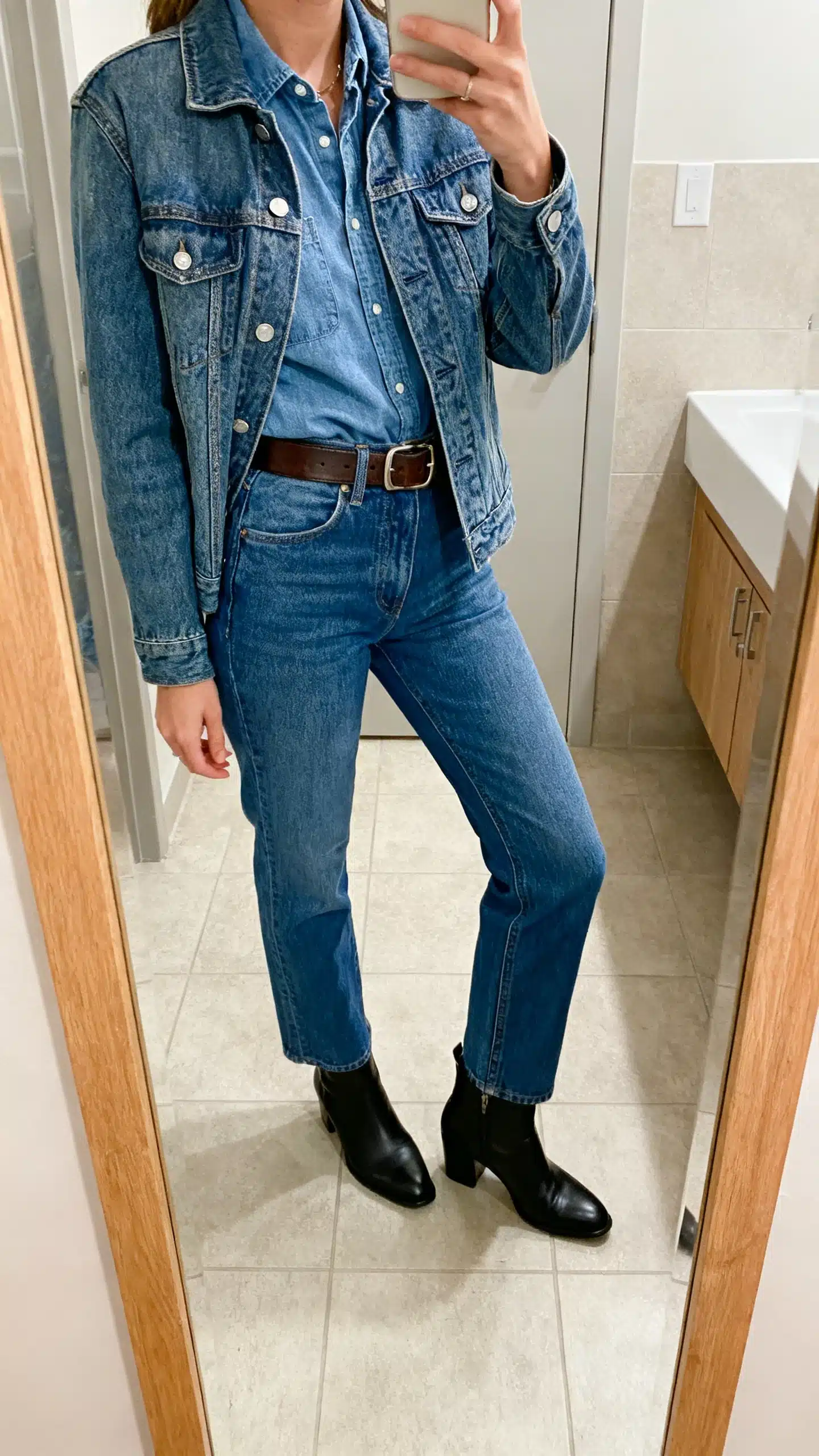 iPhone mirror selfie of a woman wearing a denim-on-denim look: medium-wash denim jacket over a chambray shirt with high-rise straight blue jeans, black ankle boots, and a leather belt, face not visible, side angle, bathroom mirror, indoor lighting, iPhone photo quality.