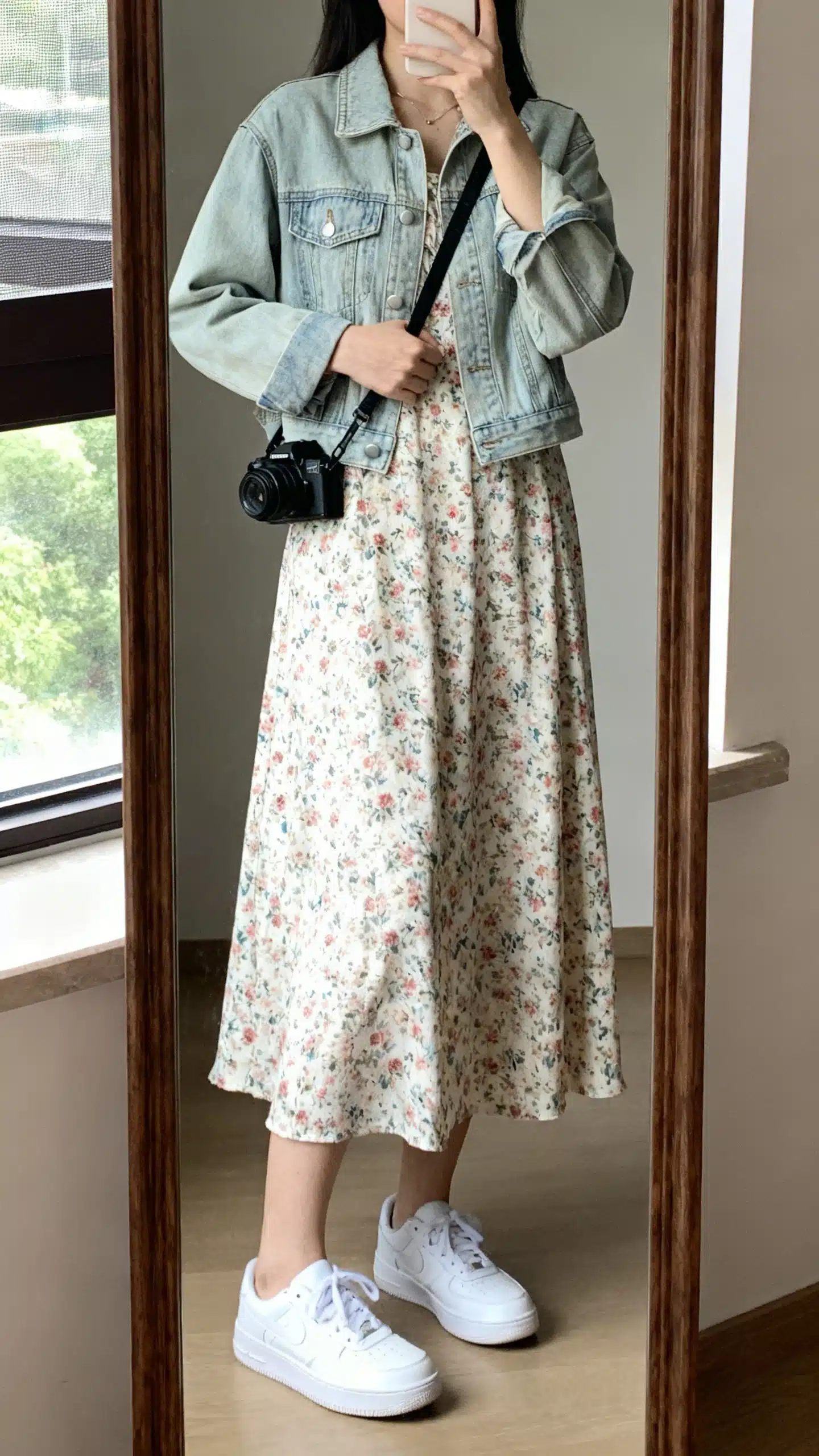 iPhone mirror selfie of a woman wearing a floral midi dress with a light-wash denim jacket, white low-top sneakers, and a crossbody camera bag, face not visible, cropped at neck, full-length mirror by window, soft natural light, iPhone photo quality.