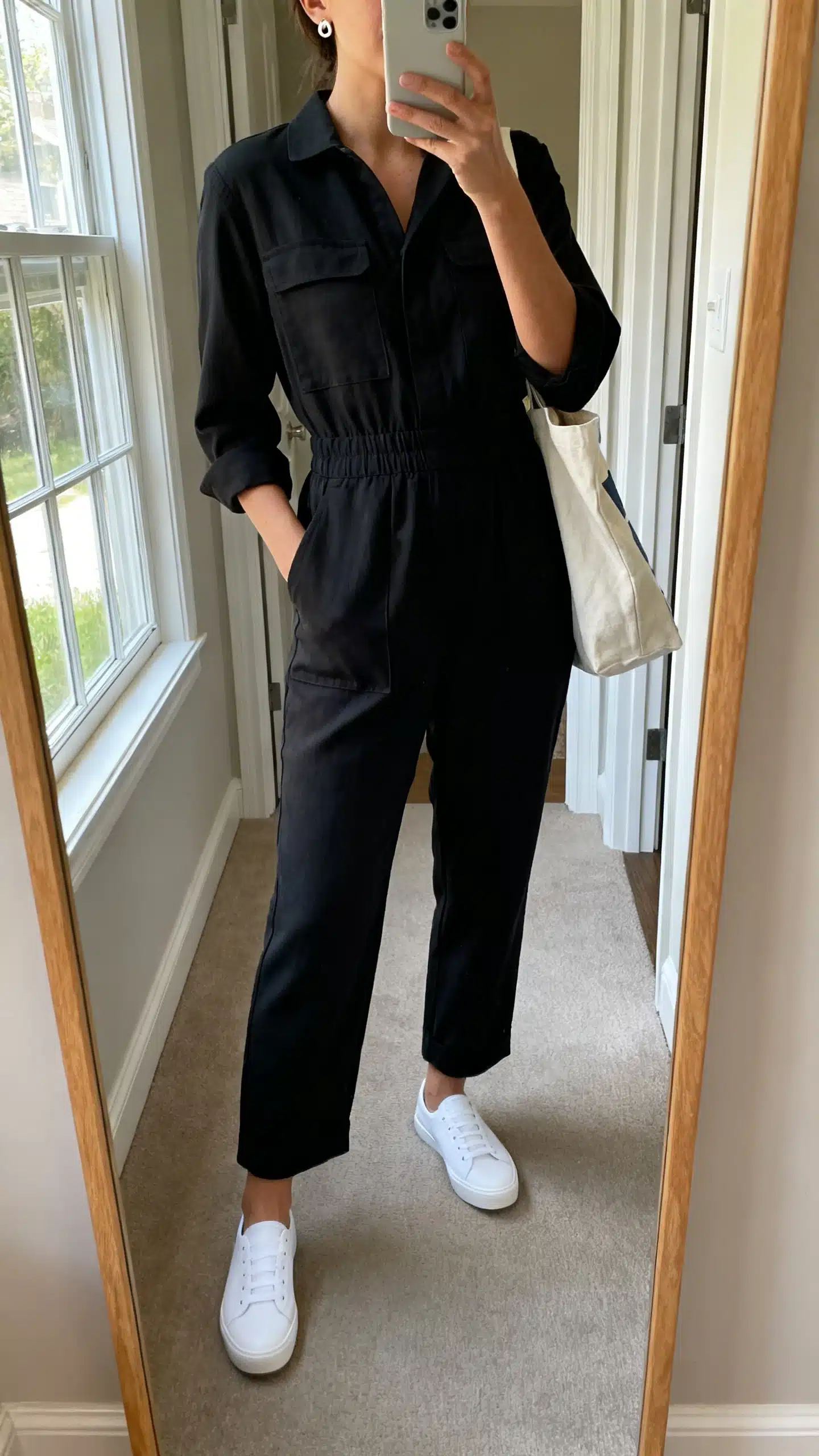 iPhone mirror selfie of a woman wearing a minimal black utility-style jumpsuit with a defined waist, white low-profile sneakers, simple stud earrings, and a tote bag, face not visible, cropped at shoulders, entryway full-length mirror, natural window light, iPhone photo quality.