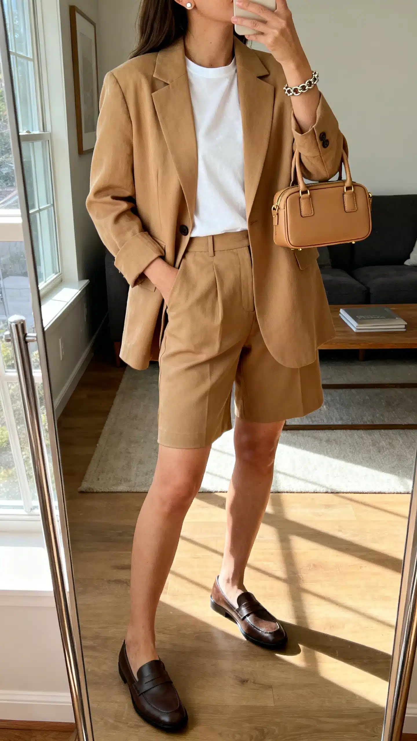 iPhone mirror selfie of a woman wearing a relaxed tan single-breasted blazer over a white tee with tailored Bermuda shorts, loafers, a structured mini tote, simple studs, and a chain bracelet, face not visible, side angle, living room full-length mirror, morning window light, iPhone photo quality.