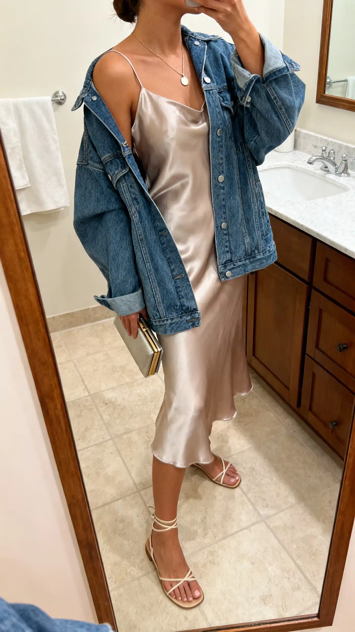 iPhone mirror selfie of a woman wearing a satin slip dress layered with an oversized denim jacket, minimalist strappy sandals, delicate pendant necklace, and a small clutch, face not visible, side angle in bathroom mirror, soft indoor lighting, iPhone photo quality.