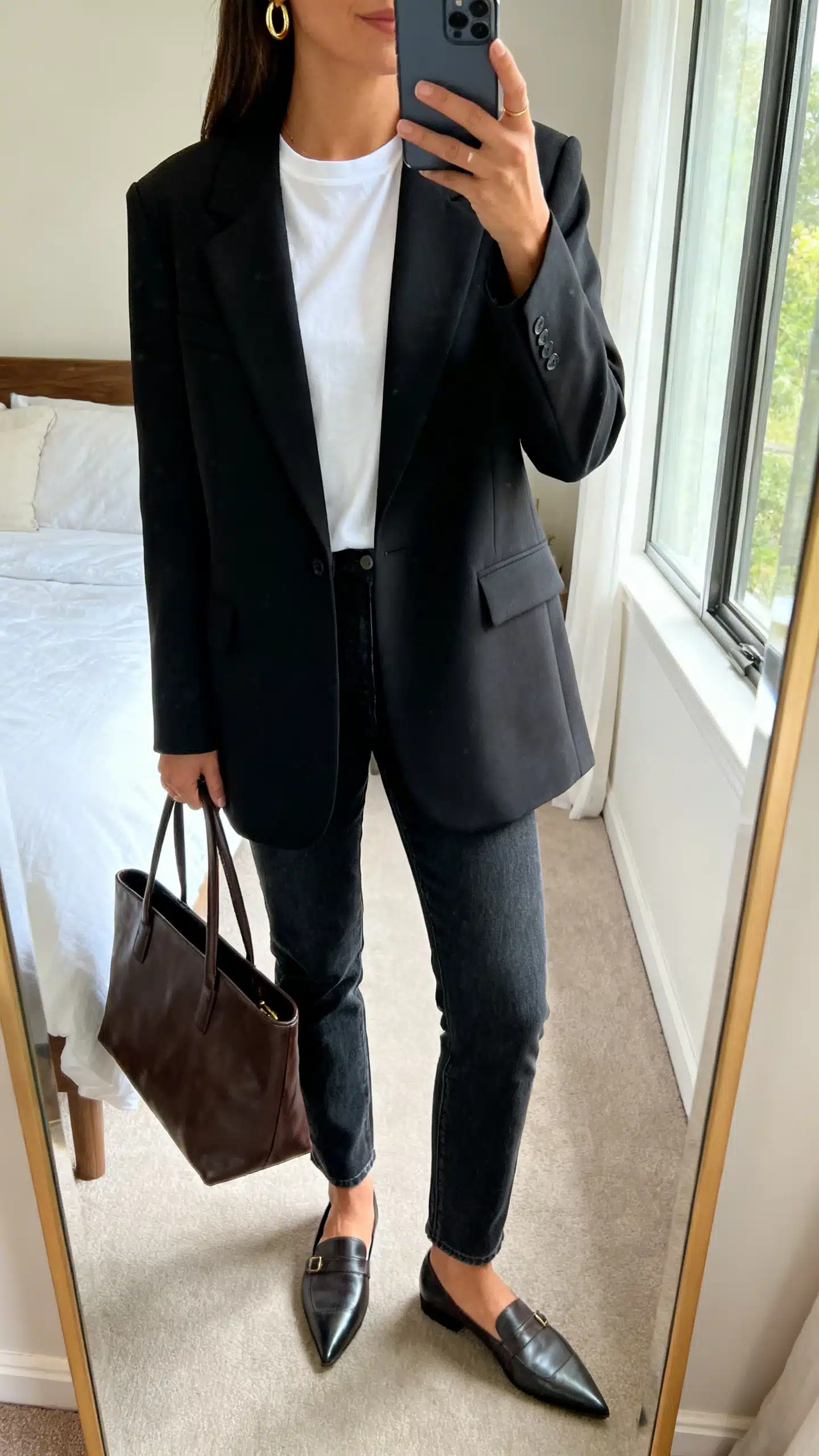 iPhone mirror selfie of a woman wearing a structured black power blazer over a white crew-neck T-shirt, slim straight jeans, pointed loafers, minimal gold hoops, and a leather tote, face not visible, cropped at neck, bedroom mirror, natural window light, iPhone photo quality.