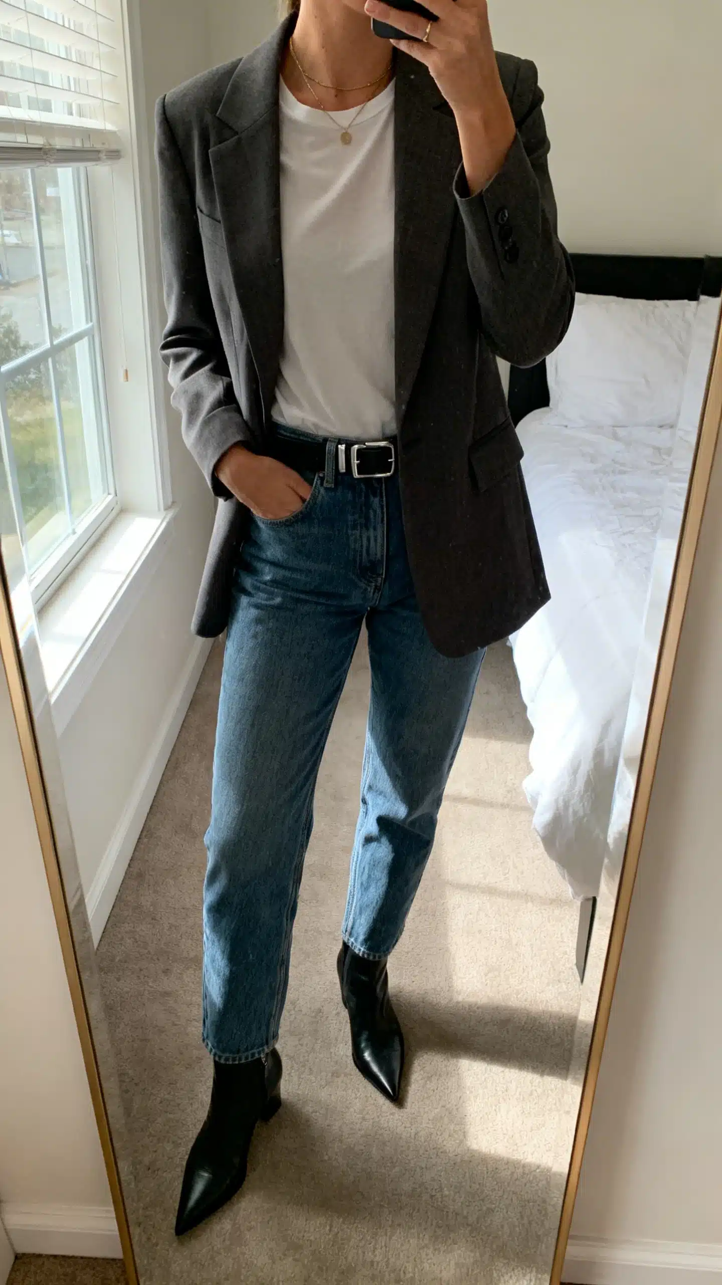 iPhone mirror selfie of a woman wearing a tailored blazer over a simple tee with straight-leg denim, sleek belt, pointed-toe ankle boots, minimal jewelry, face not visible, cropped at neck, bedroom mirror, natural morning window light, iPhone photo quality