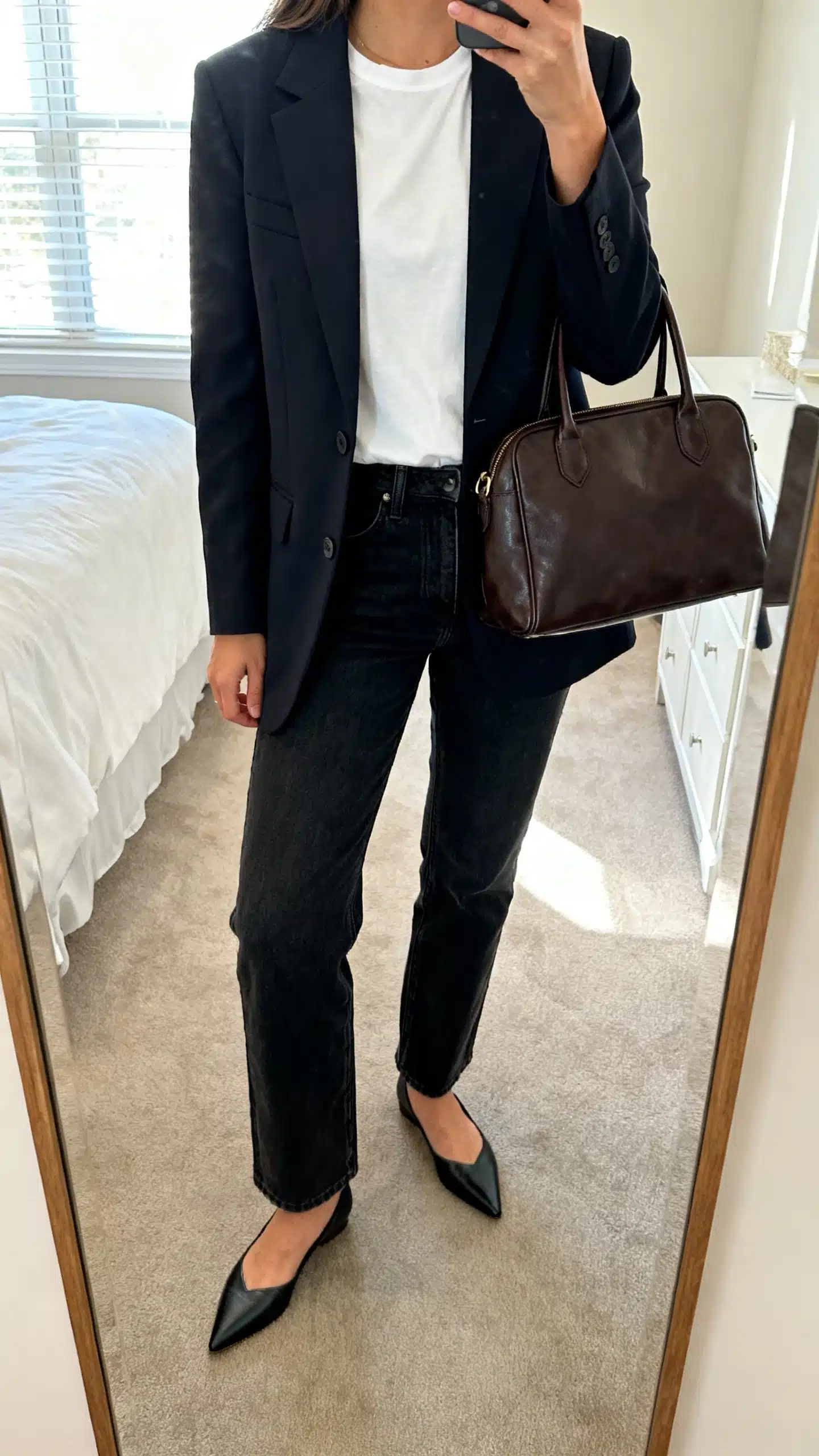 iPhone mirror selfie of a woman wearing a tailored blazer over a crisp white tee, straight-leg dark jeans, pointed flats, and a structured leather tote, face not visible, cropped at neck, bedroom mirror, natural window light, iPhone photo quality