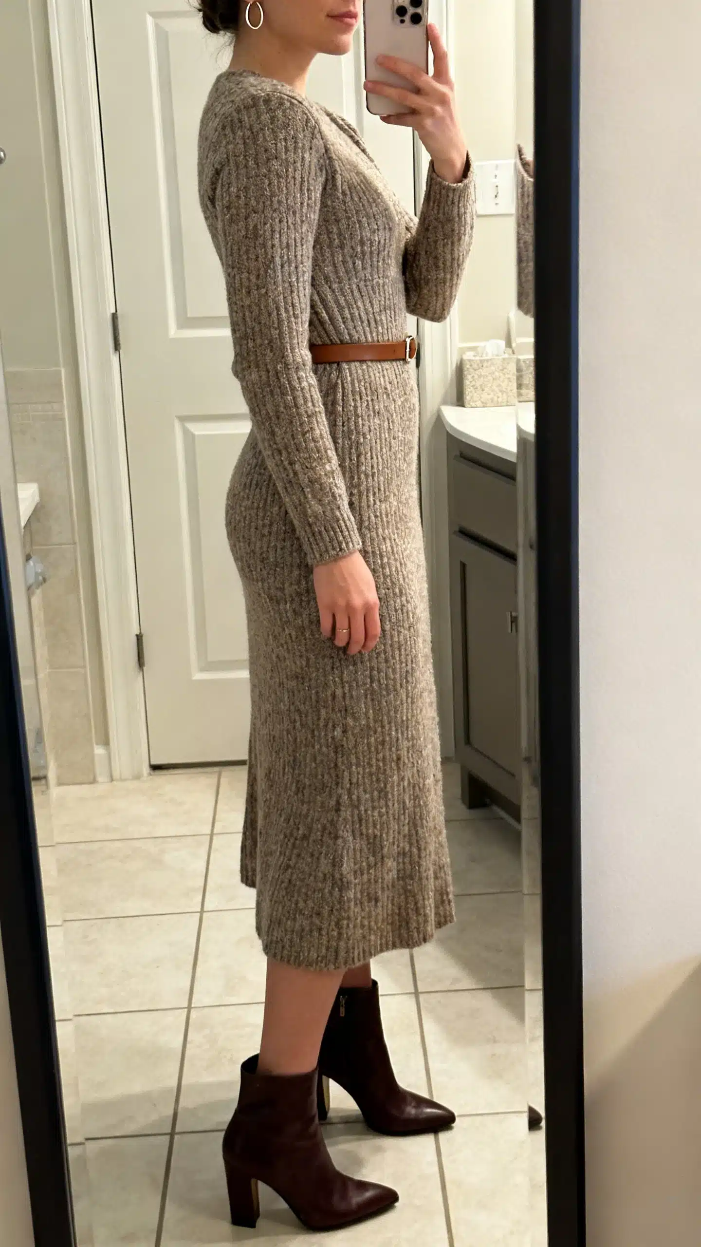 iPhone side-angle mirror selfie of a woman in a midi knit dress with a defined waist, heeled ankle boots, minimal hoop earrings, and a slim leather belt, face not visible, bathroom mirror, soft indoor lighting, iPhone photo quality