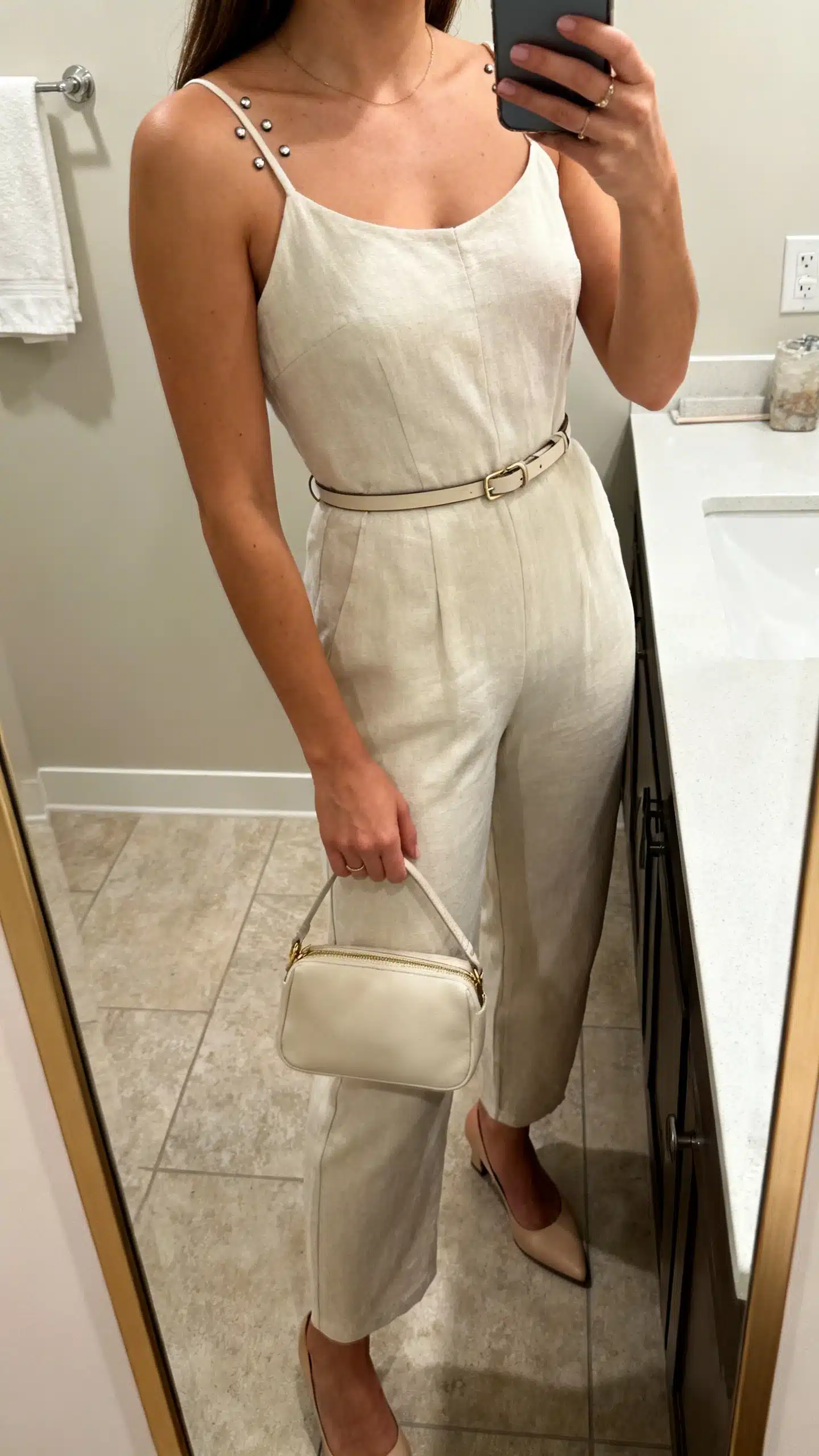 iPhone side-angle mirror selfie of a woman in a minimal jumpsuit with low heels, thin belt, understated studs, compact handbag, face not visible, cropped at shoulders, bathroom mirror, even indoor lighting, iPhone photo quality