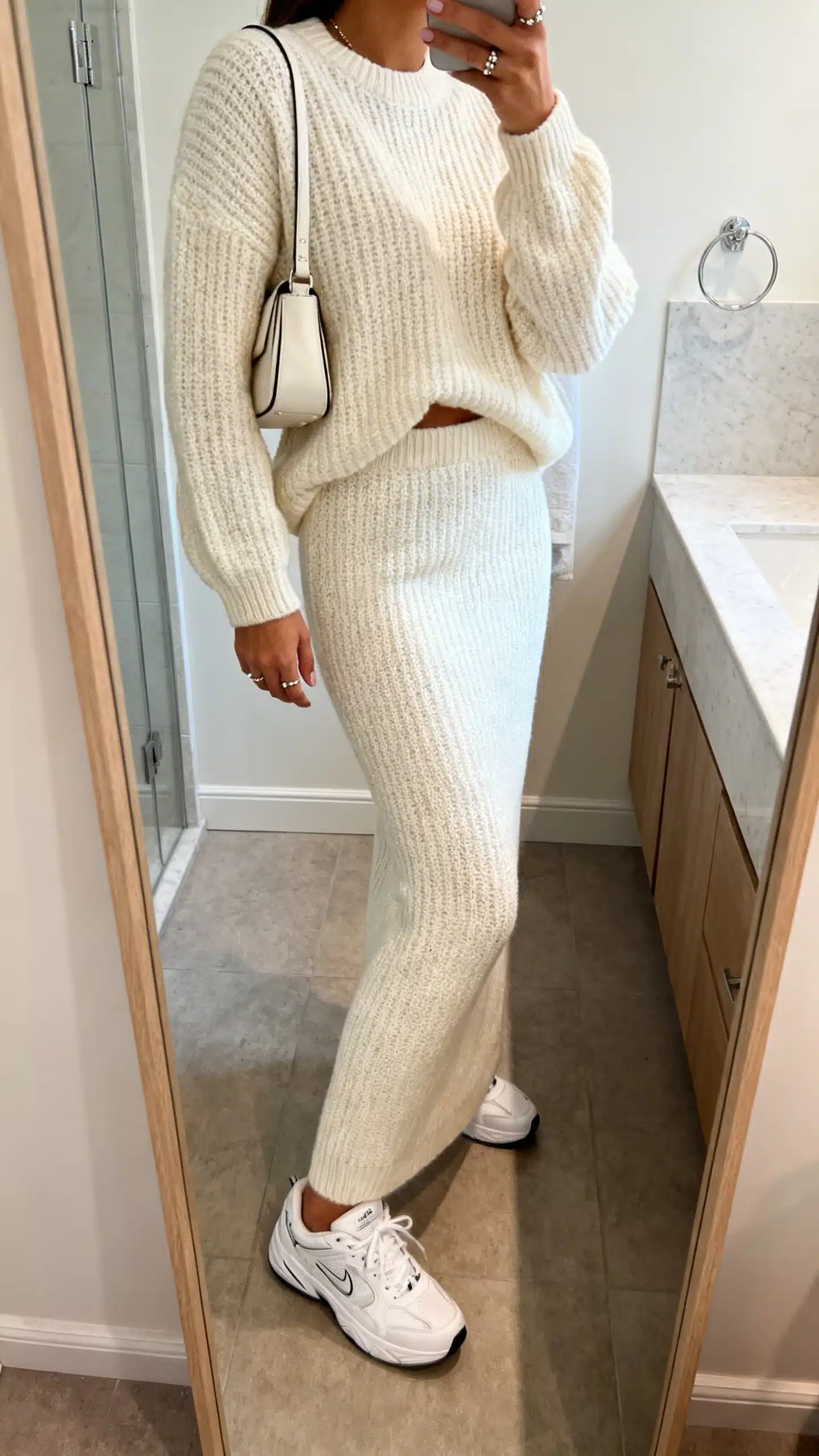 iPhone side-angle mirror selfie of a woman in a monochrome knit set (matching knit sweater and knit skirt/pants), clean white sneakers, delicate studs, small shoulder bag, face not visible, bathroom full-length mirror, soft indoor lighting, iPhone photo quality