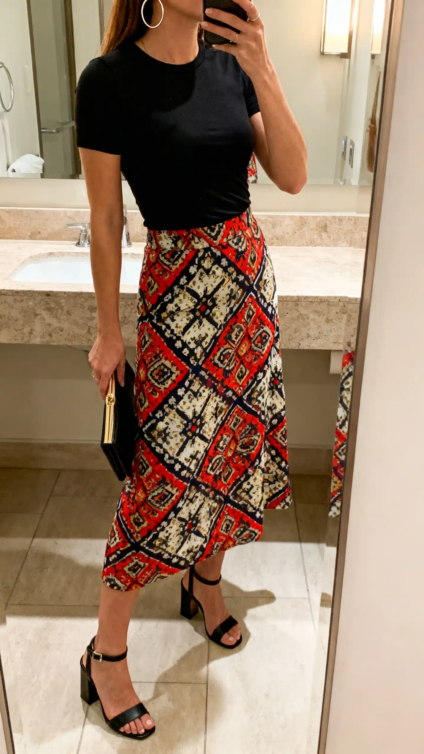 iPhone side-angle mirror selfie of a woman in a statement midi skirt (bold print) with a black fitted tee, block-heel sandals, hoop earrings, and a clutch, face not visible, bathroom mirror, indoor lighting, iPhone photo quality.