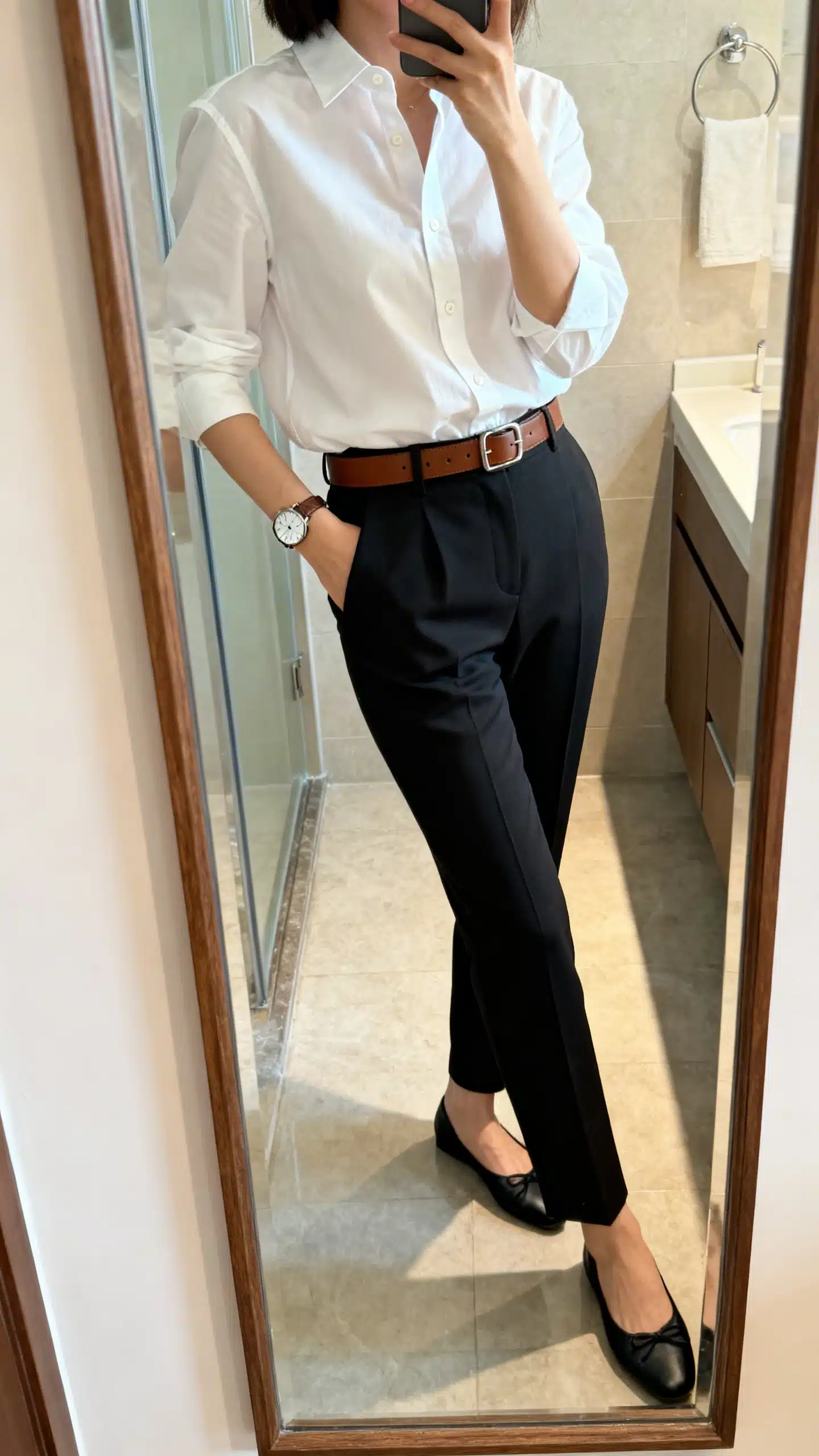 iPhone side-angle mirror selfie of a woman wearing a crisp white button-down shirt tucked into high-waisted tailored black trousers, brown leather belt, classic watch, and black ballet flats, face not visible, bathroom mirror, morning light, iPhone photo quality.