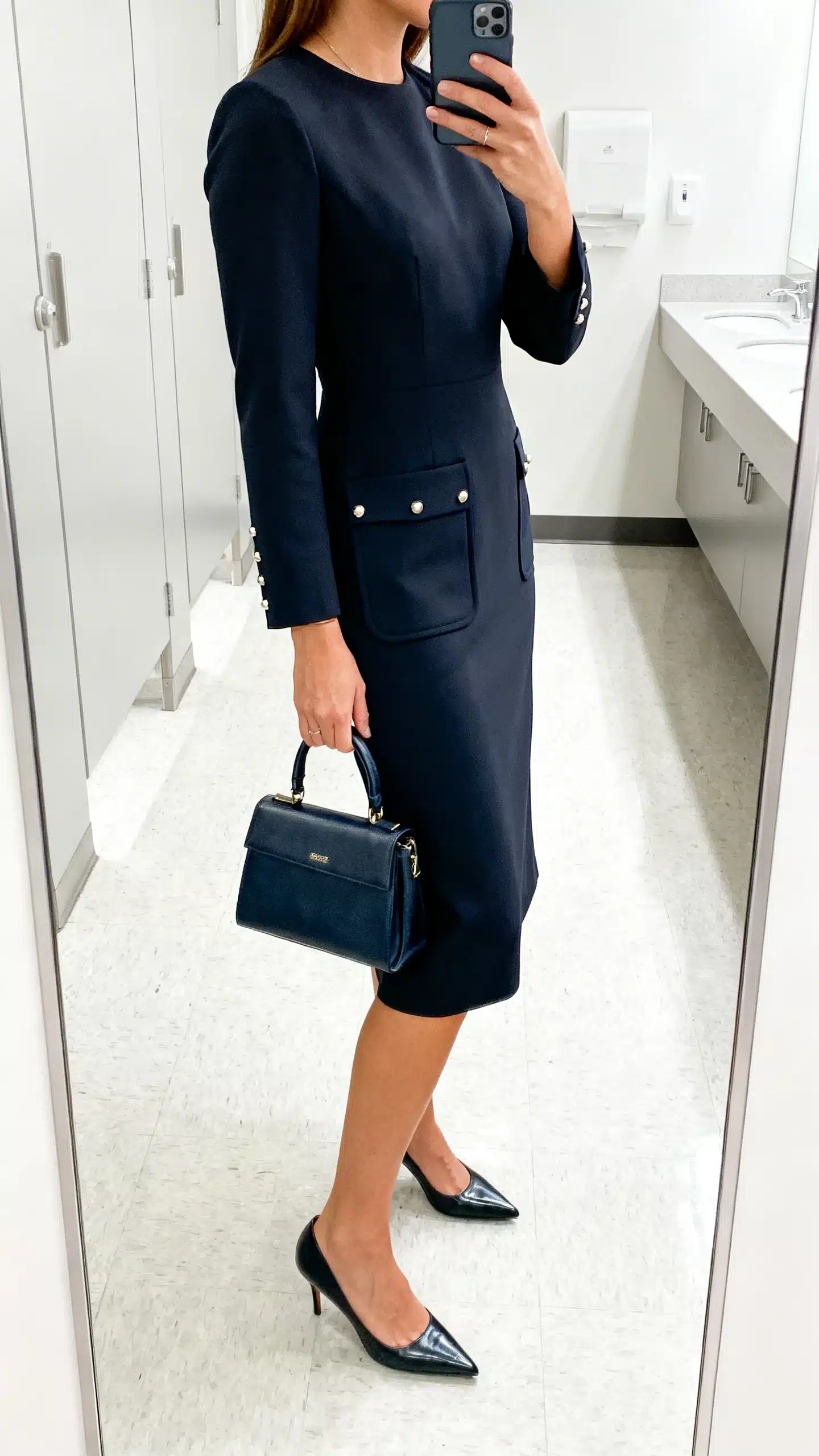 iPhone side-angle selfie of a woman in a knee-length power dress with pockets, pointed pumps, structured top-handle bag, and subtle studs, face not visible, cropped at shoulders, office restroom mirror, bright indoor lighting, iPhone photo quality
